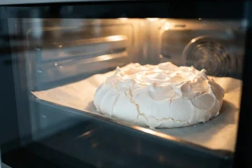 Duża biała beza Pavlova na blaszce z papierem do pieczenia wewnątrz nagrzanego piekarnika z widocznym termoobiegiem